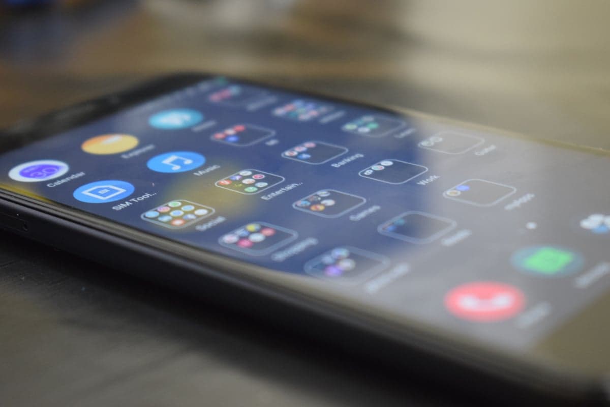 A smartphone lying on a wooden desk, its home screen filled with app icons and folders viewed at a shallow angle
