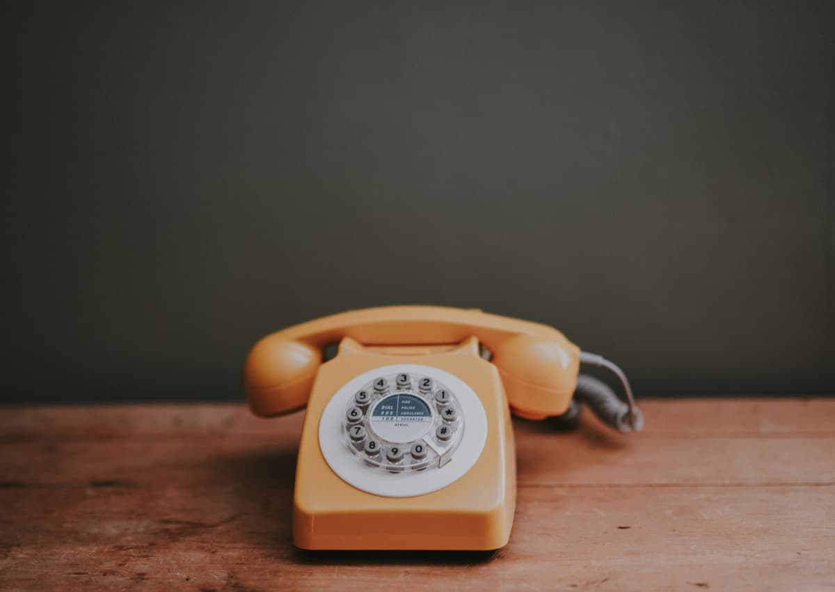 A vintage orange rotary telephone sitting on a wooden table against a dark green wall.
