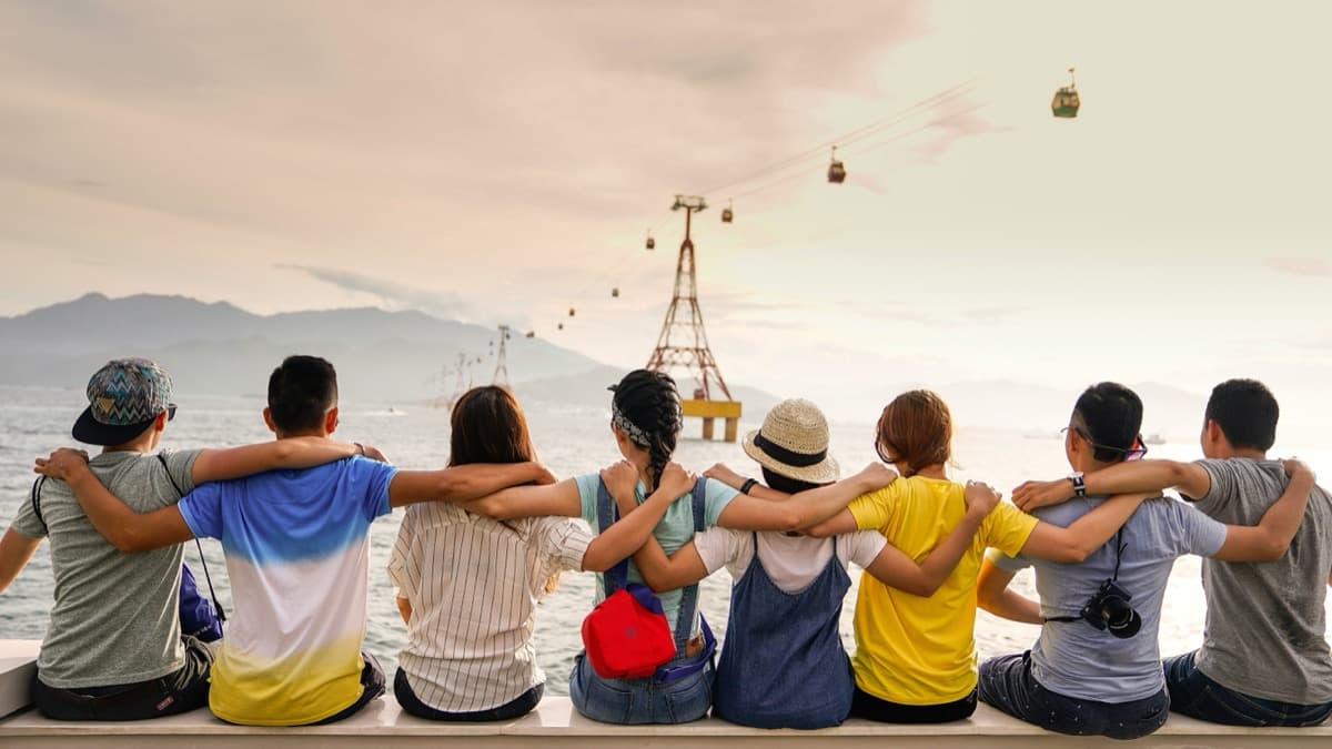 A group of seven friends photographed from behind with their arms around each other's shoulders, looking out over a hazy bay with cable cars overhead.
