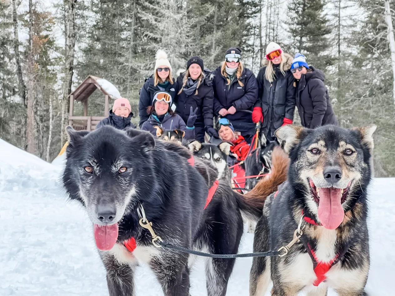 A group of women in winter gear standing behind a team of sled dogs in the snow