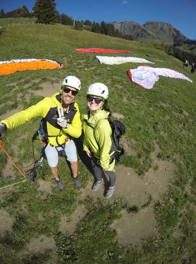 Two people in helmets and sunglasses smiling on a grassy launch slope, with brightly colored canopies laid out on the grass behind them