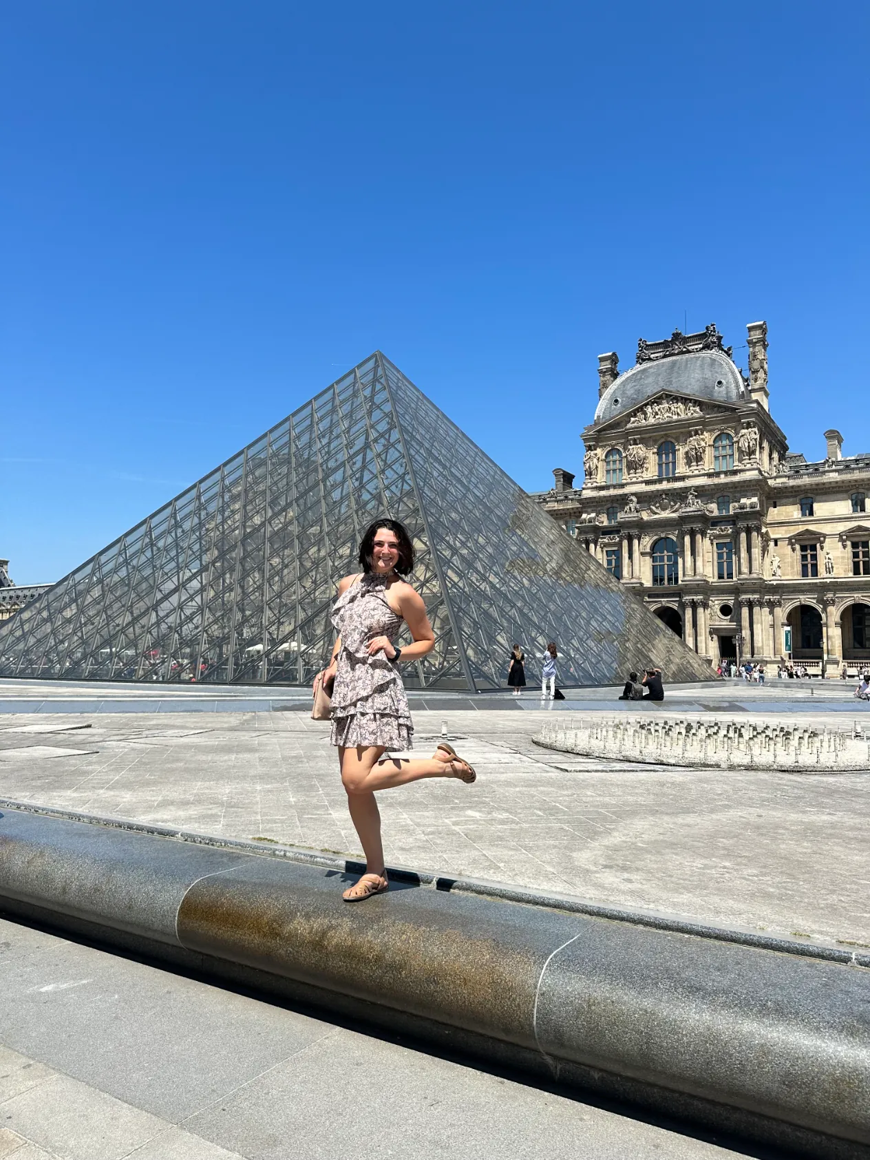 A woman posing on a stone ledge in front of the glass pyramid at the Louvre