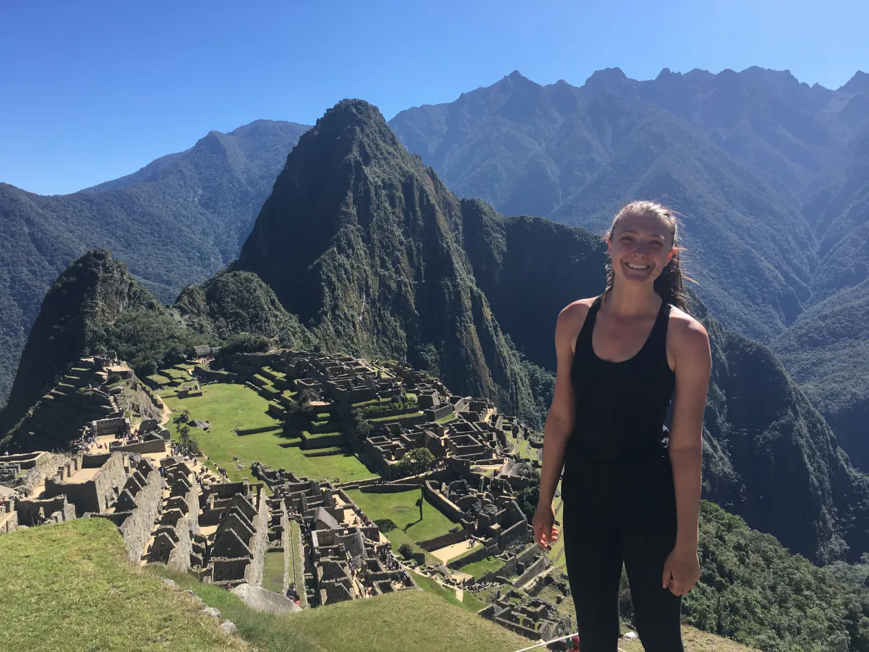 A woman standing on a grassy hillside above the stone ruins of Machu Picchu, with steep green peaks behind