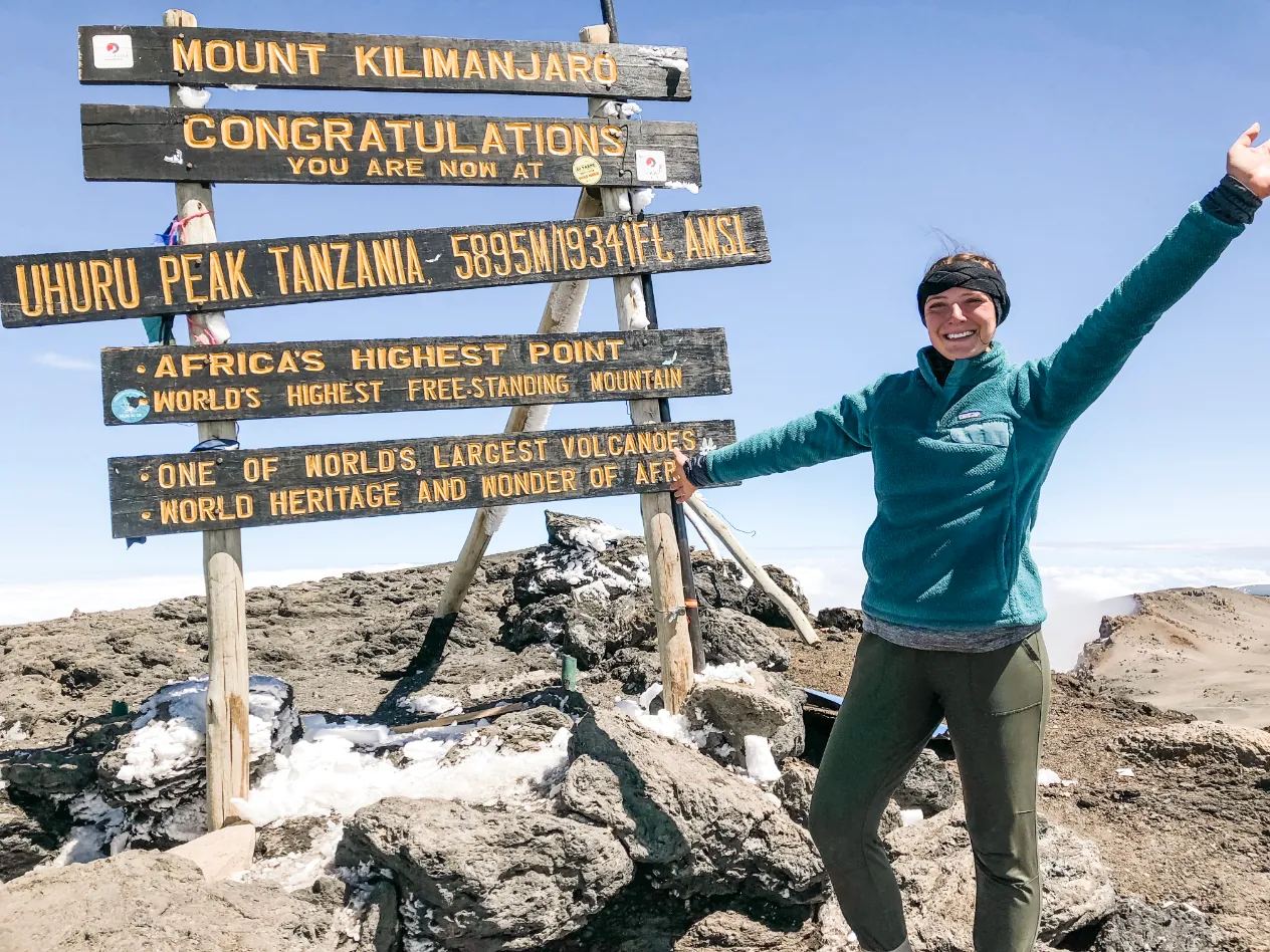 A hiker with her arms raised beside the summit sign at Uhuru Peak on Mount Kilimanjaro