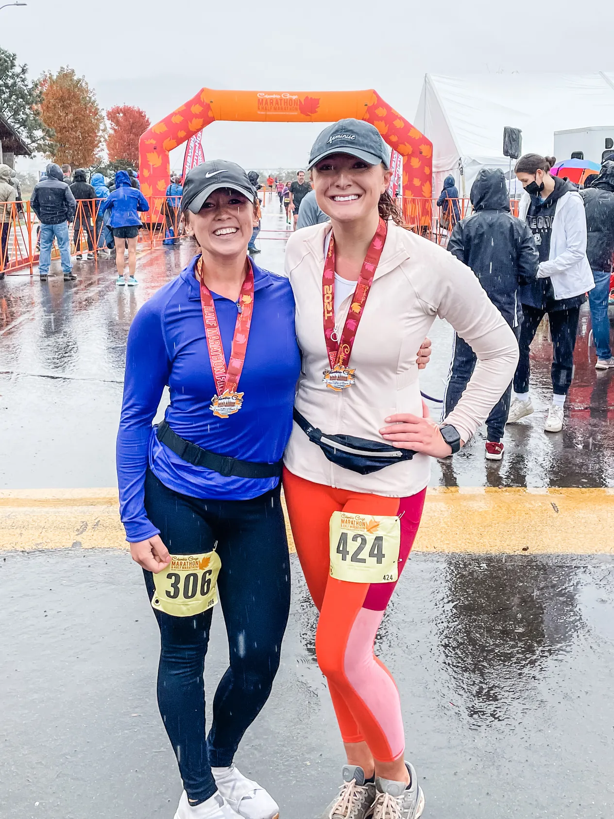 Two runners in the rain wearing finisher medals and race bibs, with an orange finish arch behind them