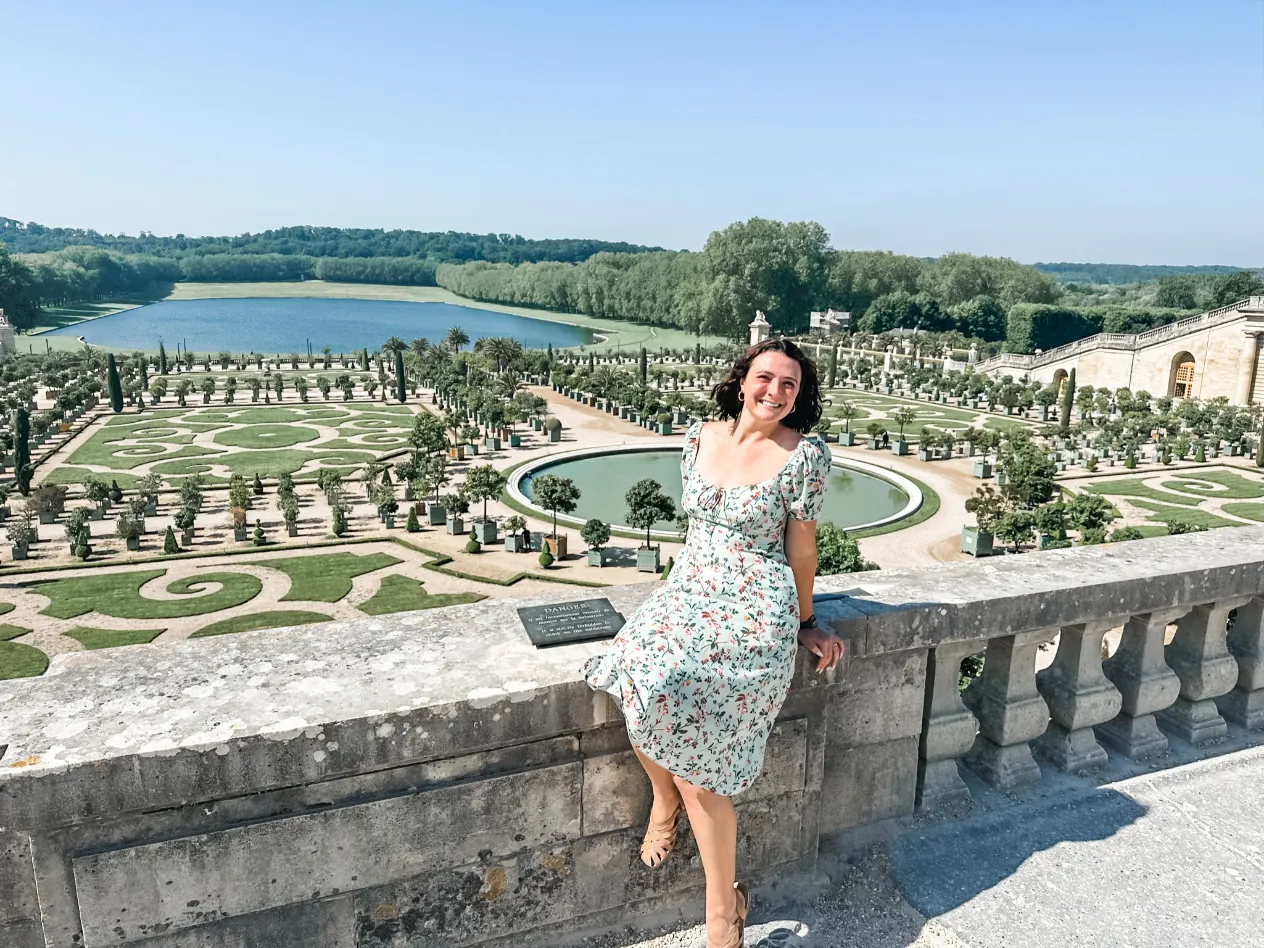 A woman in a floral dress sitting on a stone balustrade above the formal gardens at Versailles