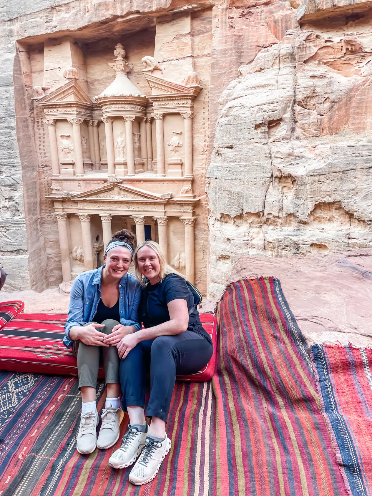 Two women sitting on striped rugs in front of the Treasury, carved into the rose-colored rock at Petra