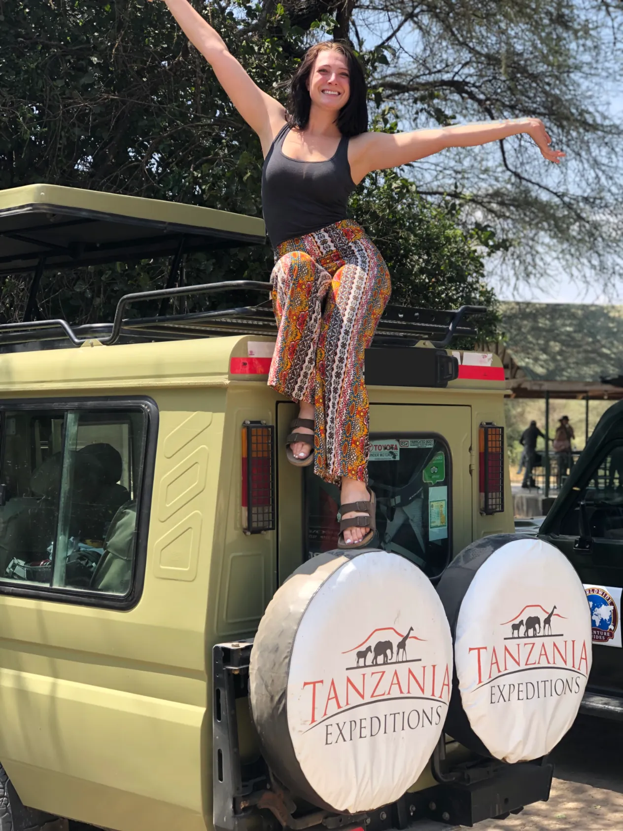 A woman with her arms out, sitting on the roof of a safari vehicle under the trees