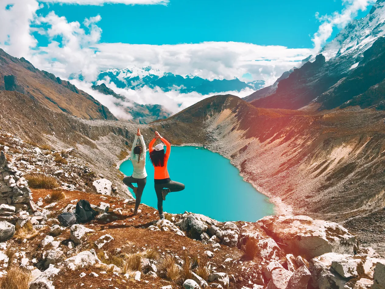 Two people in a yoga tree pose on a rocky ridge above a turquoise lake, with snowy peaks and clouds behind