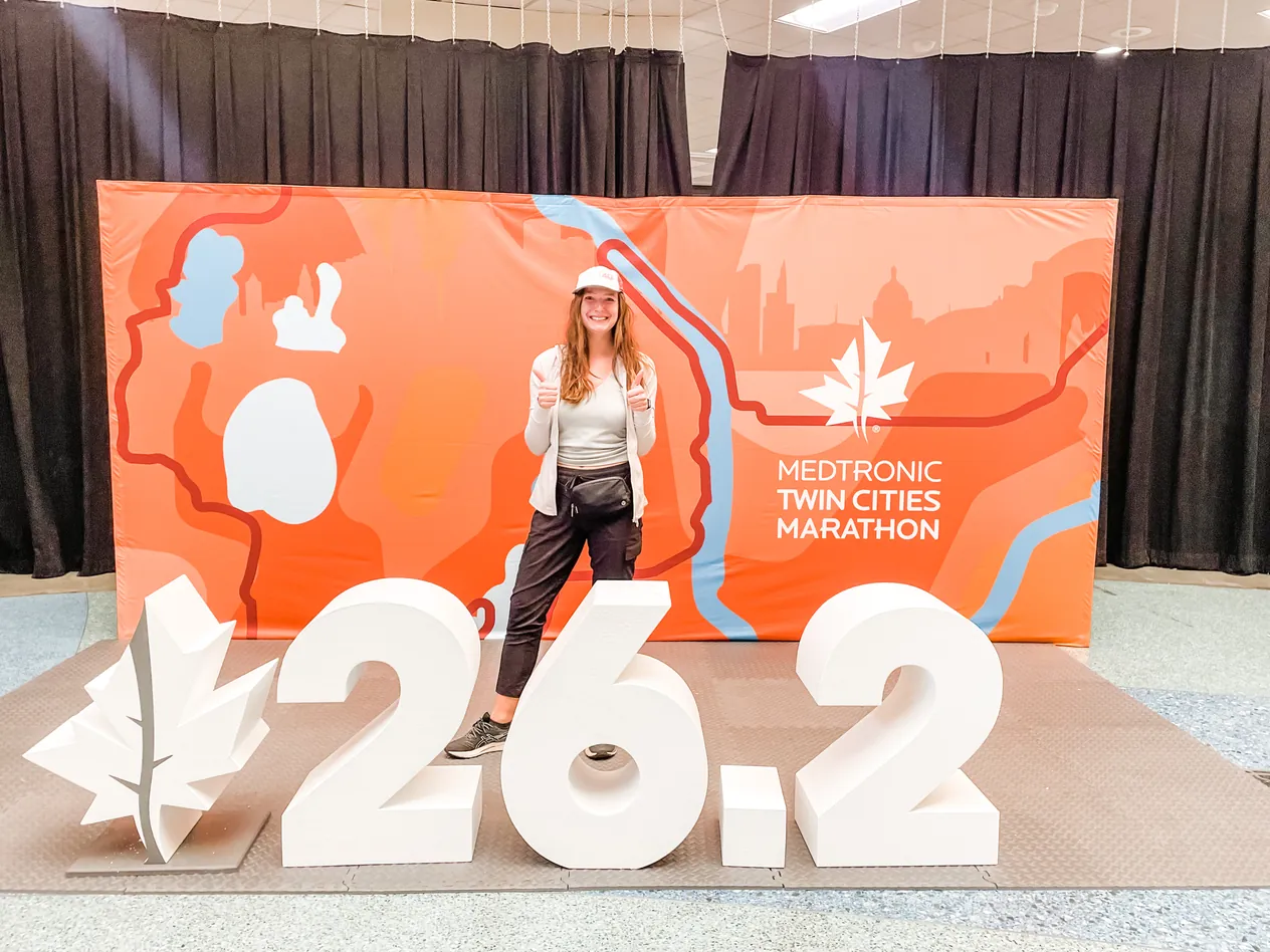 A runner giving two thumbs up in front of a giant 26.2 sign and an orange race backdrop