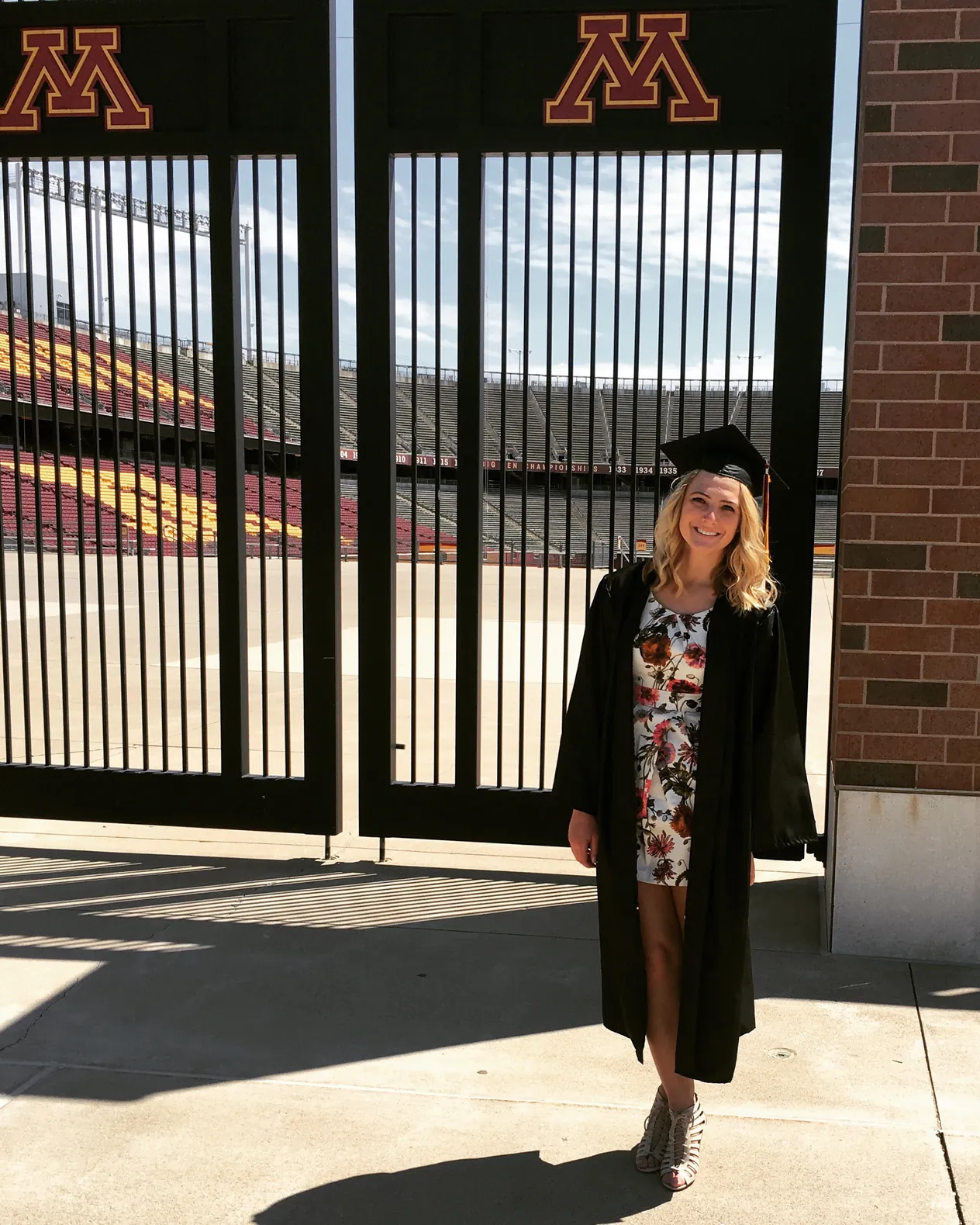 A graduate in a black gown and cap standing at the gates of the University of Minnesota football stadium